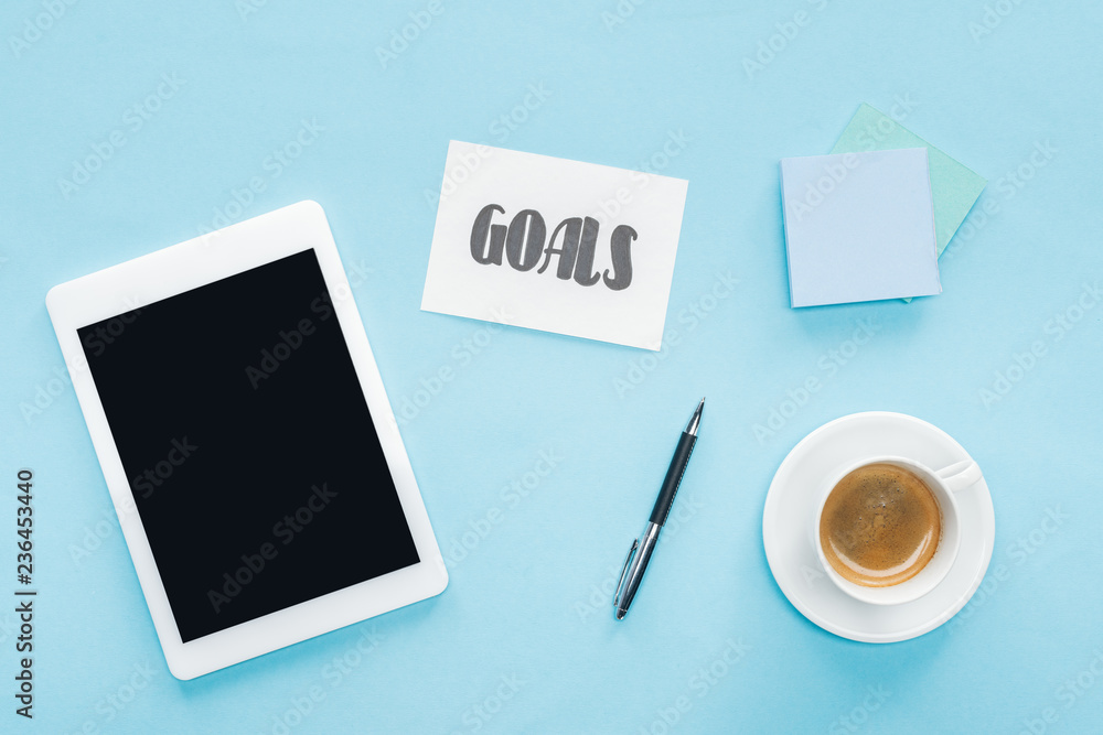 top view of digital tablet with blank screen, 'goals' lettering on card and sticky notes with blue background top view of digital tablet with blank screen, 'goals' lettering on card and sticky notes with blue background