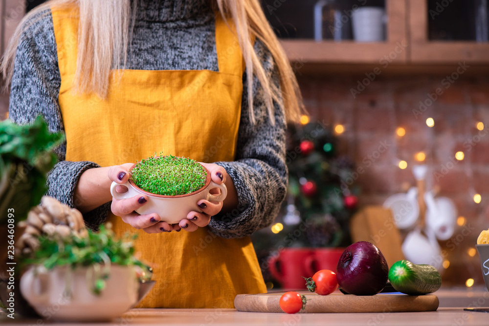 Girl with blond hair in a yellow apron in a loft kitchen, decorated with garlands, holds a bowl with microgreen/sprouts. Wooden plate with vegetables: broccoli, onions, cucumbers, microgreens etc