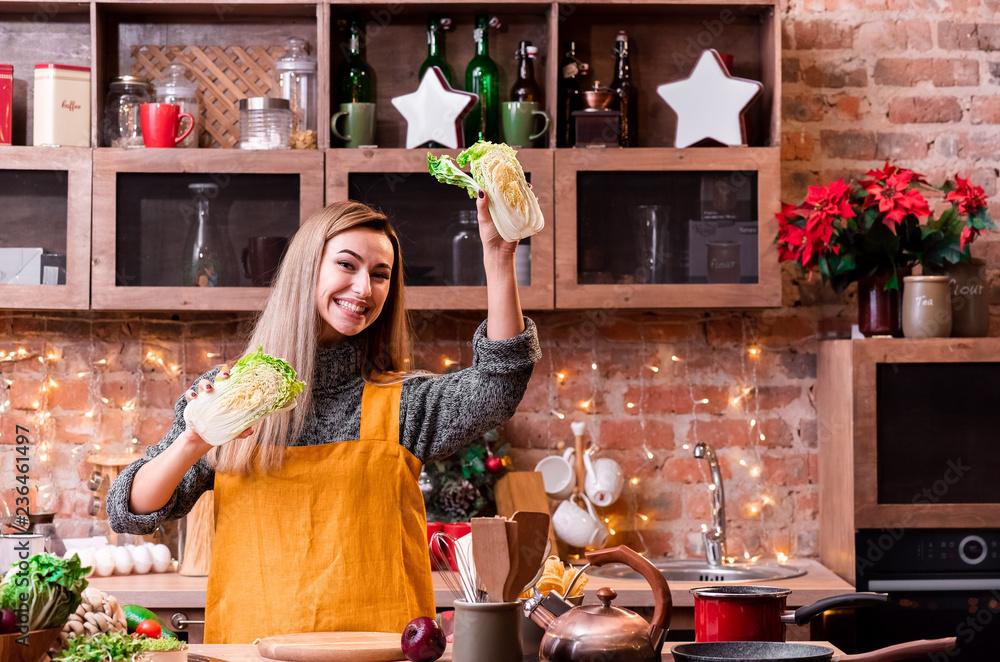 Smiled happy young girl of European appearance in a yellow apron at the loft kitchen is holding Chinese cabbage. Blonde with straight hair cooking in the loft kitchen. Space