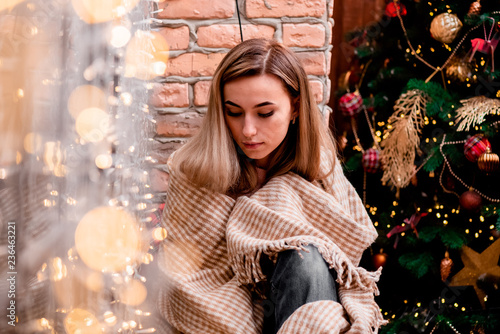 Young woman with long blond hair sitting home by the window wrapped in a blanket. Cozy loft room decorated with garlands and Christmas tree. cozy home evening. Christmas. New Year's Eve