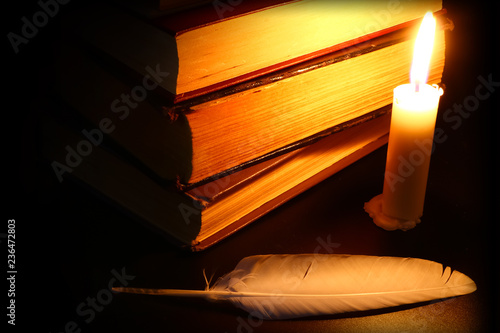 old books, burning candle and feather in the dark