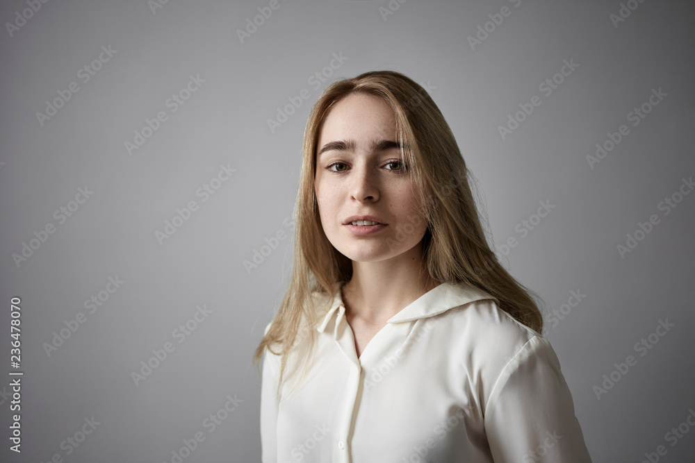 Portrait of attractive fashionable young fair haired Caucasian lady with freckles, loose hairdo and no make up posing in studio wearing white shirt at gray copyspace wall, looking at camera Portrait of attractive fashionable young fair haired Caucasian lady with freckles, loose hairdo and no make up posing in studio wearing white shirt at gray copyspace wall, looking at camera