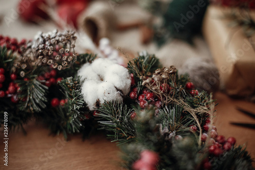 Details of rustic Christmas wreath, cotton close up. Fir branches with red berries,pine cones,ribbon, cinnamon on rustic wood. Atmospheric moody image at holiday workshop