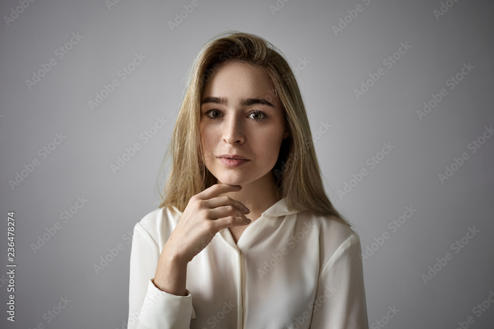People, femininity, beauty, style and fashion concept. Horizontal shot of attractive fair haired young Caucasian woman with loose hair and freckles tocuhing her chin, having serious thoughtful look People, femininity, beauty, style and fashion concept. Horizontal shot of attractive fair haired young Caucasian woman with loose hair and freckles tocuhing her chin, having serious thoughtful look