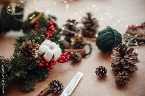 Fir branches, wreath, red berries, pine cones, thread, scissors, cinnamon, cotton, lights on rustic wooden background. Details for making christmas wreath at workshop. Atmospheric image