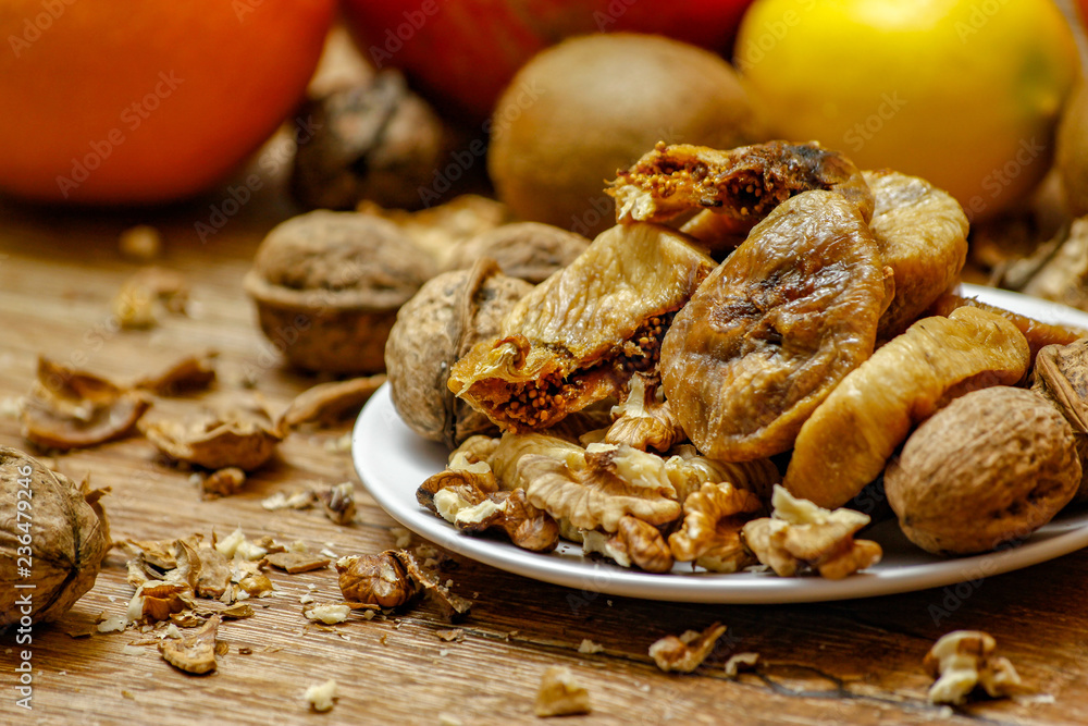 Dried fig and whole and cracked walnuts on white ceramic plate. Wooden table and various citrus in the background. Dried fig and whole and cracked walnuts on white ceramic plate. Wooden table and various citrus in the background.
