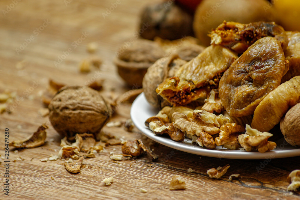 Dried fig and whole and cracked walnuts on white ceramic plate. Wooden table and various citrus in the background. Dried fig and whole and cracked walnuts on white ceramic plate. Wooden table and various citrus in the background.