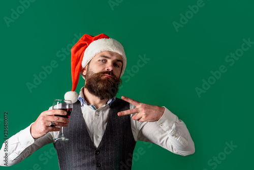 Confident successful man in Santa hat holds glass of wine, showing victory or peace gesture. Feeling relaxed&chill. Man in waistcoat drink glass of red wine. Santa man tasting red wine. New year party