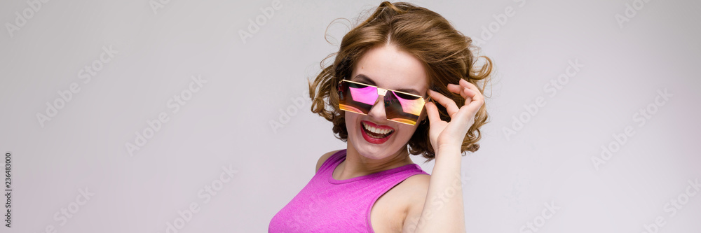 Naklejka premium Charming young girl in pink dress on gray background. Cheerful girl in square glasses. The girl adjusts glasses