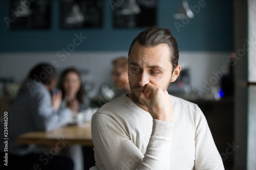 Upset millennial outsider feel offended lack company, young outcast guy suffer from discrimination, jealous of friends hang out together in cafe, envious male loner depressed sit alone in coffeeshop