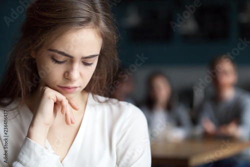 Upset young girl sitting alone in cafe offended at friends joke, sad millennial woman suffer from low self-esteem, depressed female outcast feeling outsider hanging out with company in coffeeshop photo