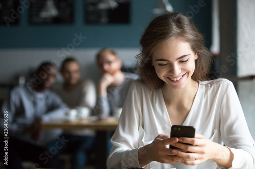 Pretty young girl sit in cafe using smartphone laughing because of flirting students from behind table, happy female smiling feeling attention from males talking to her. Love from first sight concept