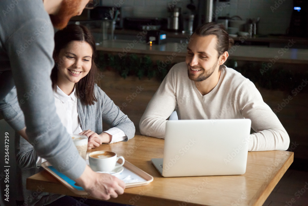 Waiter give order to smiling millennial couple sitting at coffeeshop table with laptop, young cafe worker bring cappuccino and latte to happy coffeehouse visitors, server serving cups with hot drinks Waiter give order to smiling millennial couple sitting at coffeeshop table with laptop, young cafe worker bring cappuccino and latte to happy coffeehouse visitors, server serving cups with hot drinks