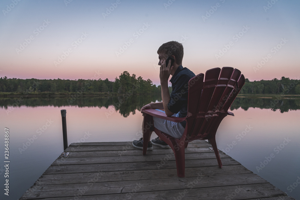 Young man on his cell phone while watching the sunset from a chair on a dock.