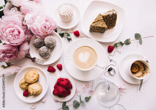 Delicious morning breakfast table essentials: fresh cappuccino coffee, marshmallow, cookies, chocolate, red raspberries, glass of water and tender pink blossoming peony bouquet on the white background