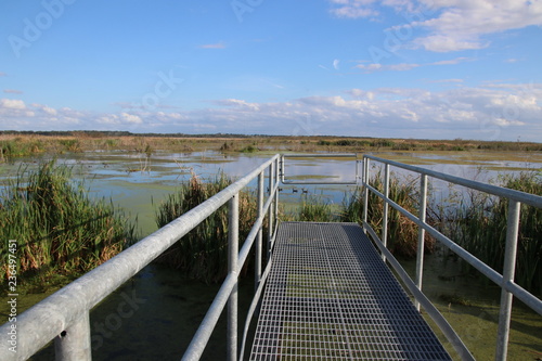 Perspective photography metal bridge, dock or pier walkway leading out over blue water lake, pond or river with blue sky horizon.