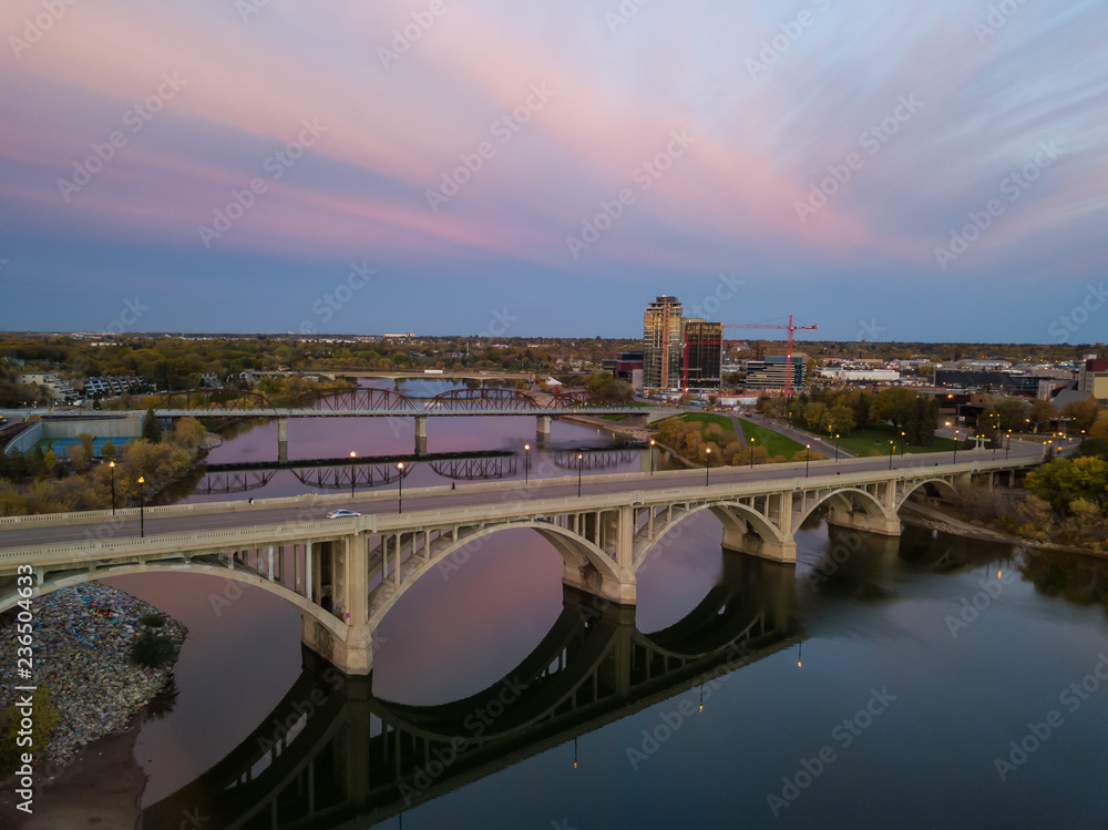 Naklejka premium Aerial view of a bridge going over Saskatchewan River during a vibrant sunrise in the Fall Season. Taken in Saskatoon, SK, Canada.