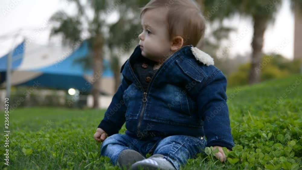 Portrait of cute stylish smiling toddler boy sitting on the grass in park outdoors. Positive smiling handsome blonde boy wearing blue jeans
