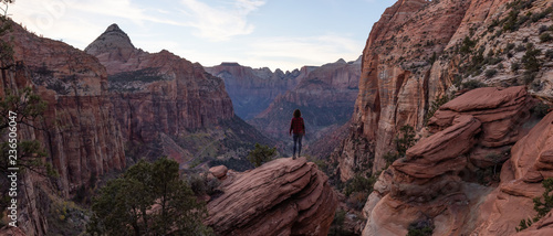 Adventurous Girl at the edge of a cliff is looking at a beautiful landscape view in the Canyon during a vibrant sunset. Taken in Zion National Park, Utah, United States.