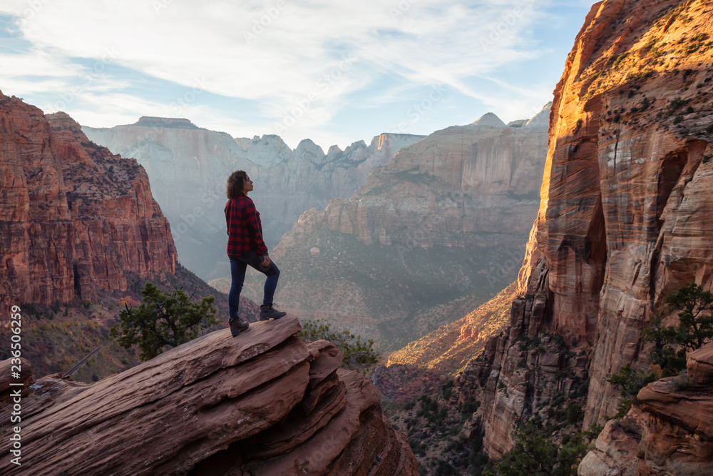 Fototapeta premium Adventurous Girl at the edge of a cliff is looking at a beautiful landscape view in the Canyon during a vibrant sunset. Taken in Zion National Park, Utah, United States.