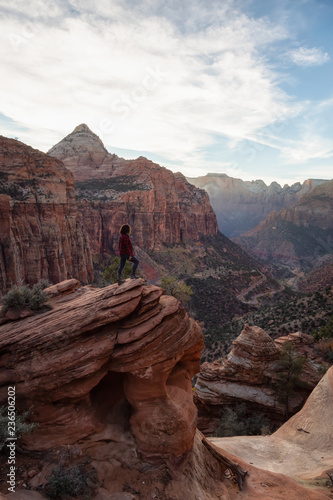 Adventurous Girl at the edge of a cliff is looking at a beautiful landscape view in the Canyon during a vibrant sunset. Taken in Zion National Park, Utah, United States.