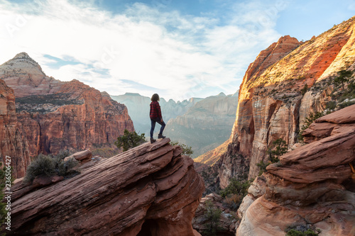 Adventurous Woman at the edge of a cliff is looking at a beautiful landscape view in the Canyon during a vibrant sunset. Taken in Zion National Park  Utah  United States.