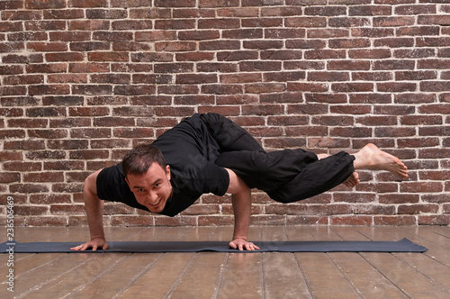 Sporty attractive young man working out, yoga, pilates, fitness training, asana Dwi pada kaundiniasana over brick wall background. Studio shot. Full length photo