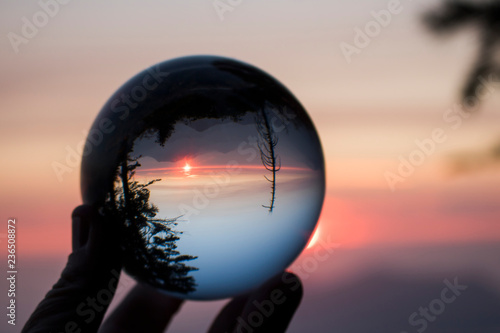 Fototapeta Naklejka Na Ścianę i Meble -  Glass Ball in Fingertips Captures Reflection of Sunset over Sierra Nevada Mountain Range with Forest Trees in Silhouette