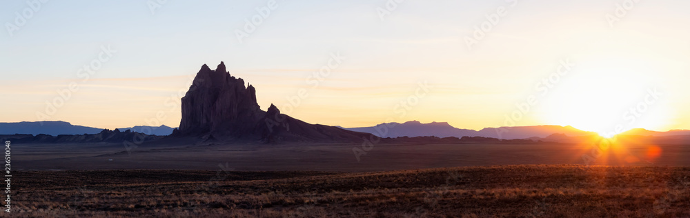 Striking panoramic landscape view of a dry desert with a mountain peak in the background during a vibrant sunset. Taken at Shiprock, New Mexico, United States.
