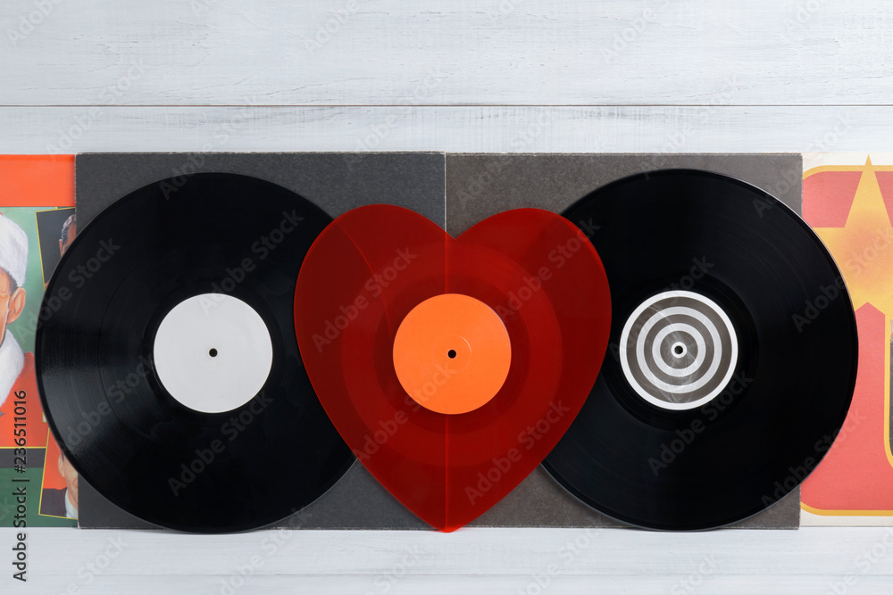 A red heart-shaped vinyl record between two black vinyl records for a  turntable with a gray covers white wooden background of their boards.  Listening to Christmas songs. Valentine's Day, Christmas Stock Photo
