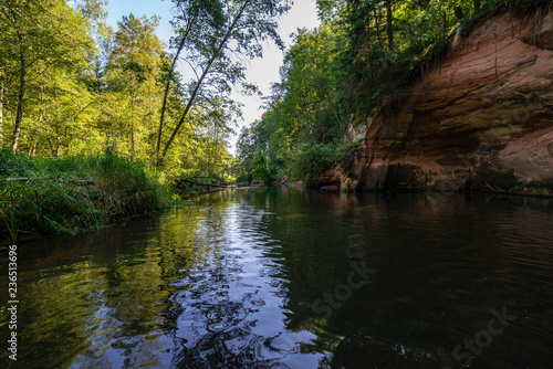 summer day on water in calm river enclosed in forests with sandstone cliffs and dry wood © Martins Vanags