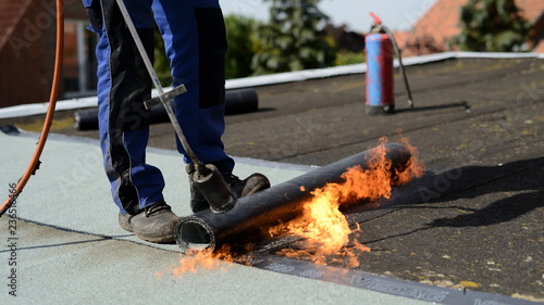 Dachdecker auf Flachdach Baustelle in der Industrie macht Dach neu: Isolierter Flachdach Geselle schweißt industriell Flachdach Dachpappe in Handarbeit mit offener Flamme auf flaches Dach