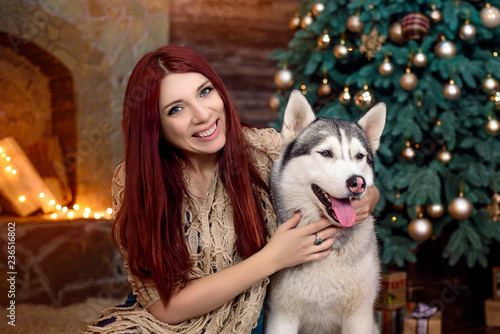 Happy woman sitting on floor and hugging two husky dogs near christmas tree. Happy winter holiday concept. © hohmochka