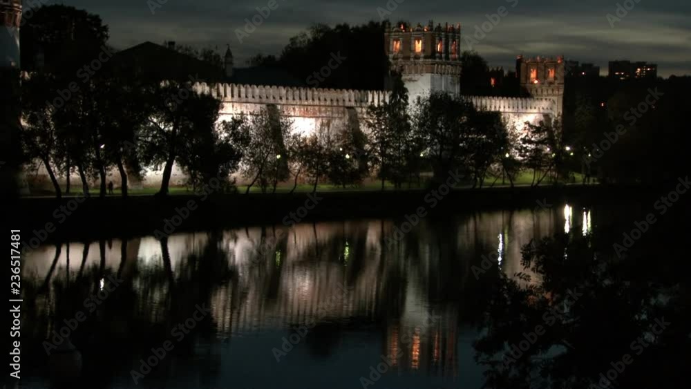 People walk near Novodevichy monastery on shore of pond at night.