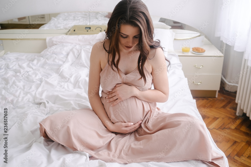 Young woman holds her pregnant belly and sitting on the bed 