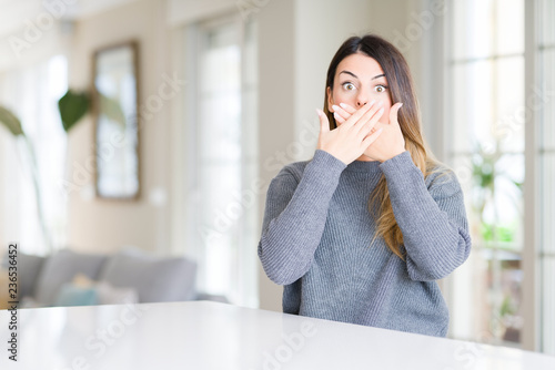 Young beautiful woman wearing winter sweater at home shocked covering mouth with hands for mistake. Secret concept.