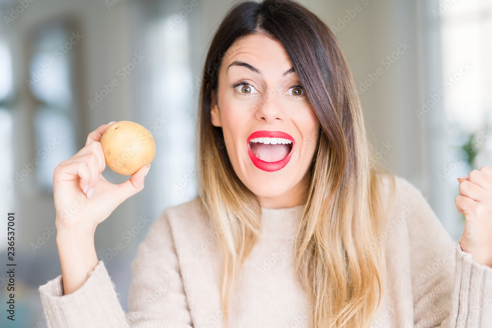 Young beautiful woman holding fresh potato at home screaming proud and celebrating victory and success very excited, cheering emotion Young beautiful woman holding fresh potato at home screaming proud and celebrating victory and success very excited, cheering emotion