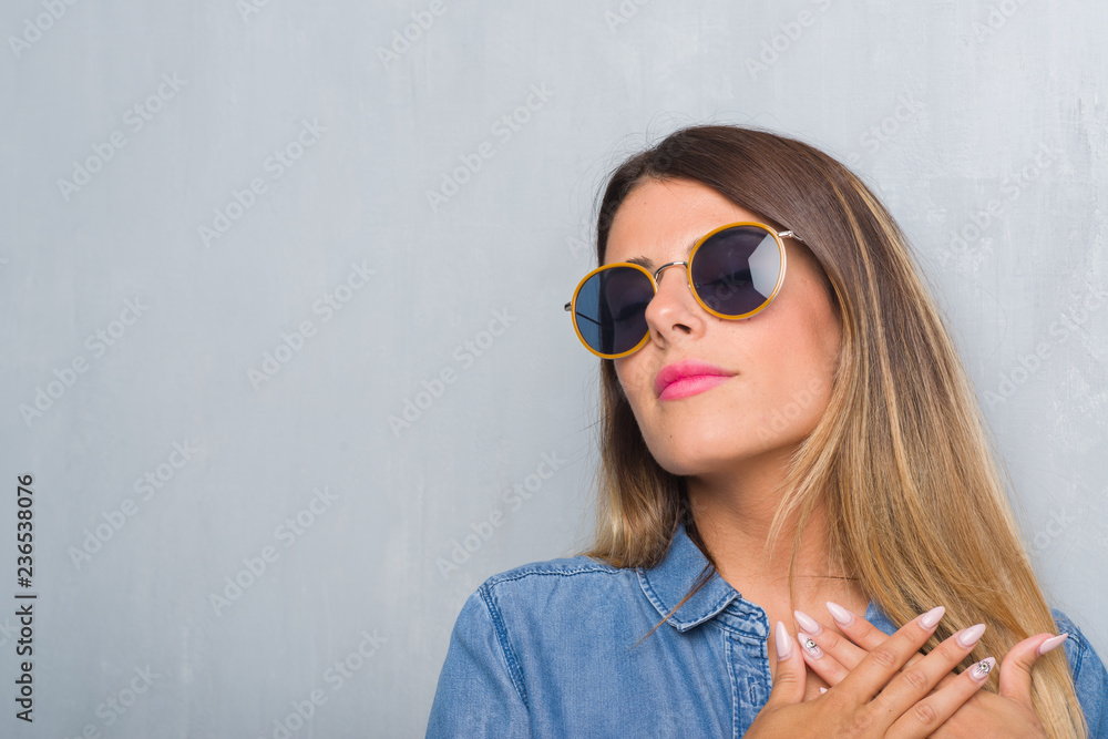 Young adult woman over grunge grey wall wearing retro sunglasses smiling with hands on chest with closed eyes and grateful gesture on face. Health concept. Young adult woman over grunge grey wall wearing retro sunglasses smiling with hands on chest with closed eyes and grateful gesture on face. Health concept.