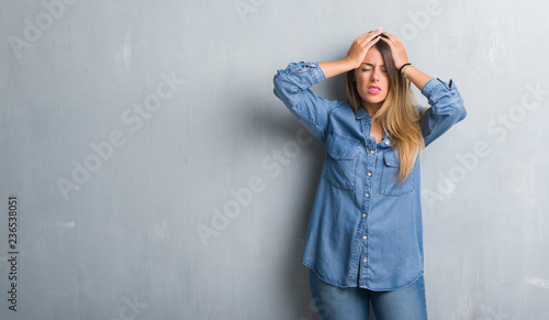 Young adult woman over grunge grey wall wearing denim outfit suffering from headache desperate and stressed because pain and migraine. Hands on head.