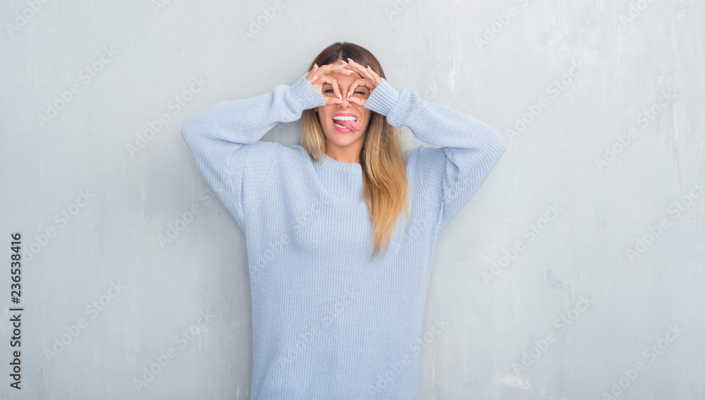 Young adult woman over grey grunge wall wearing winter outfit doing ok gesture like binoculars sticking tongue out, eyes looking through fingers. Crazy expression. Young adult woman over grey grunge wall wearing winter outfit doing ok gesture like binoculars sticking tongue out, eyes looking through fingers. Crazy expression.