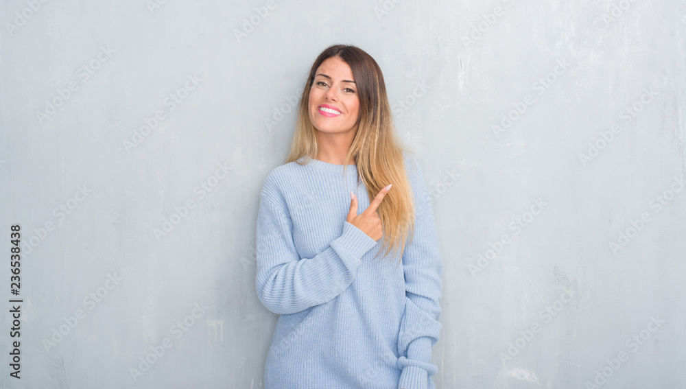 Young adult woman over grey grunge wall wearing winter outfit cheerful with a smile of face pointing with hand and finger up to the side with happy and natural expression on face Young adult woman over grey grunge wall wearing winter outfit cheerful with a smile of face pointing with hand and finger up to the side with happy and natural expression on face