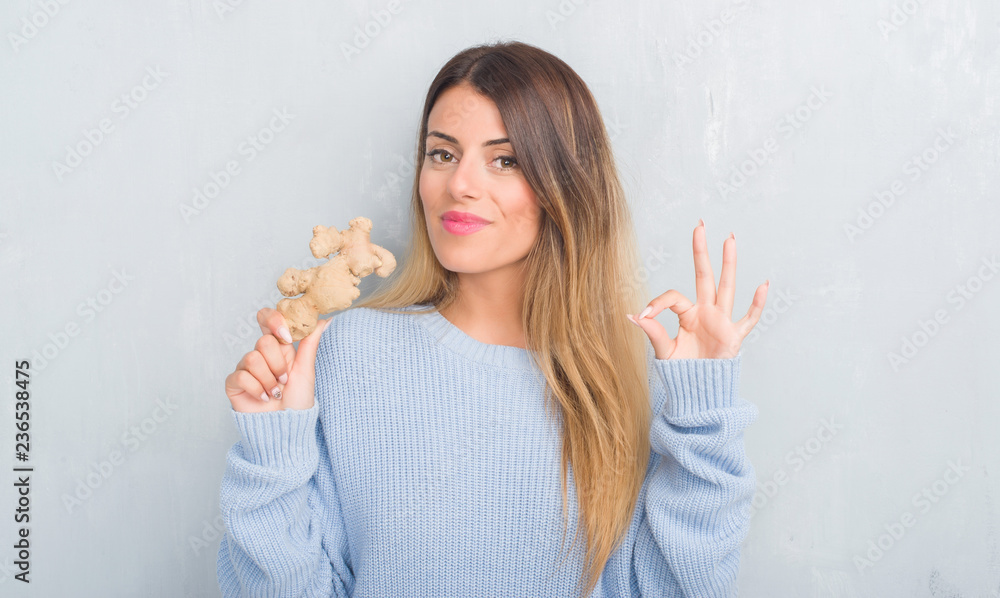 Young adult woman over grey grunge wall holding fresh ginger vegatable doing ok sign with fingers, excellent symbol Young adult woman over grey grunge wall holding fresh ginger vegatable doing ok sign with fingers, excellent symbol