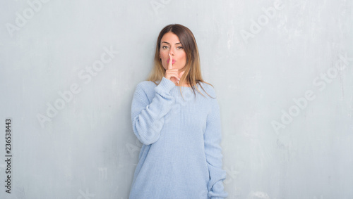 Young adult woman over grey grunge wall wearing winter outfit asking to be quiet with finger on lips. Silence and secret concept. © Krakenimages.com