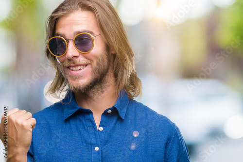 Young handsome man with long hair wearing sunglasses over isolated background smiling with happy face looking and pointing to the side with thumb up.