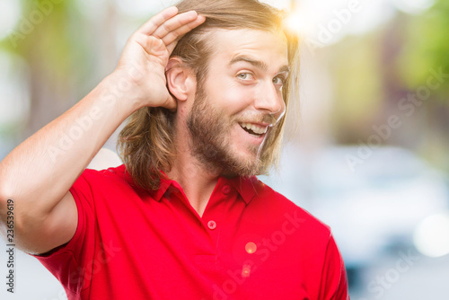 Young handsome man with long hair over isolated background smiling with hand over ear listening an hearing to rumor or gossip. Deafness concept.