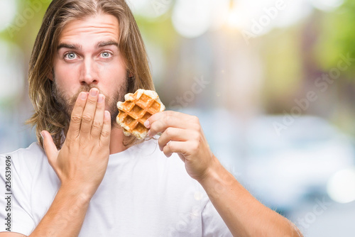 Young handsome man with long hair over isolated background eating waffle cover mouth with hand shocked with shame for mistake, expression of fear, scared in silence, secret concept