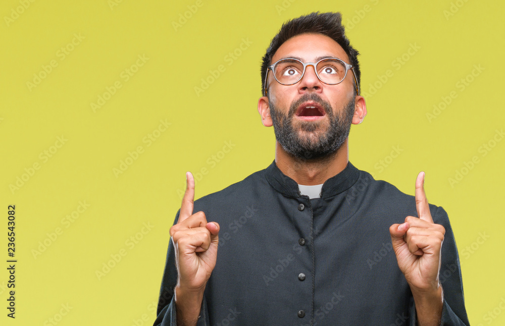 Adult hispanic catholic priest man over isolated background amazed and surprised looking up and pointing with fingers and raised arms. Adult hispanic catholic priest man over isolated background amazed and surprised looking up and pointing with fingers and raised arms.