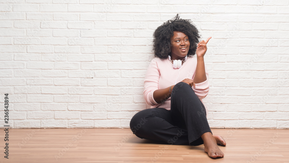 Young african american woman sitting on the floor wearing headphones with a big smile on face, pointing with hand and finger to the side looking at the camera. Young african american woman sitting on the floor wearing headphones with a big smile on face, pointing with hand and finger to the side looking at the camera.