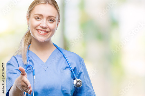 Young blonde surgeon doctor woman over isolated background smiling friendly offering handshake as greeting and welcoming. Successful business.