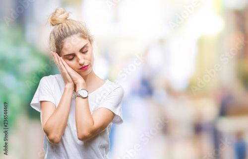 Young beautiful blonde woman wearing white t-shirt over isolated background sleeping tired dreaming and posing with hands together while smiling with closed eyes.